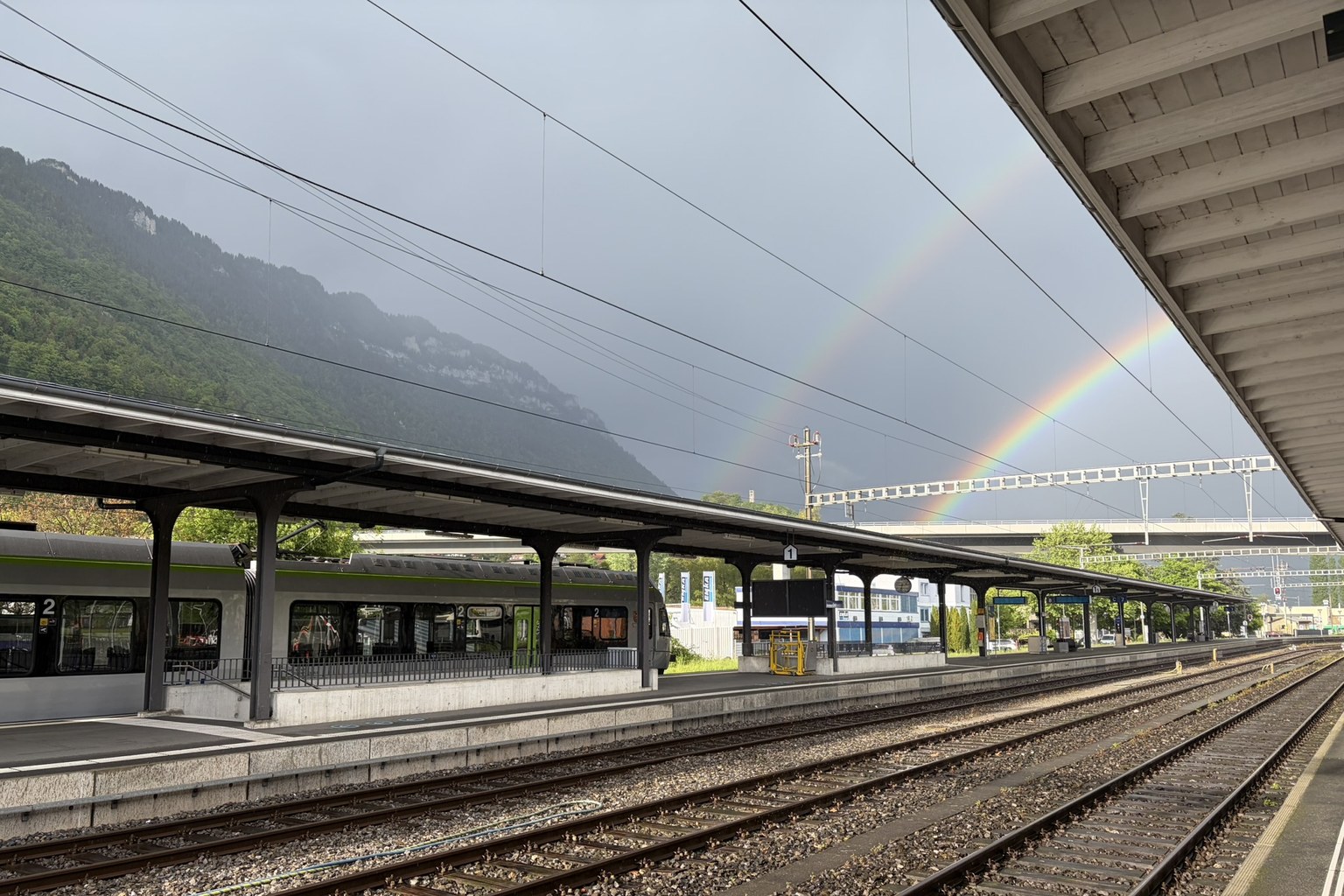 瑞士火車站月台，雙彩虹橫跨陰暗天空，前景為延伸向遠方的鐵軌，左側停靠一列區間列車